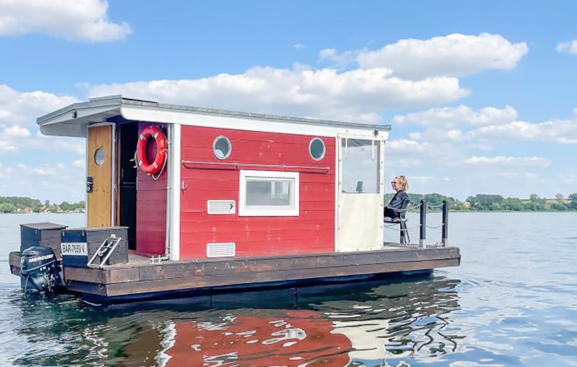 Floß unterwegs Rotes schwimmendes Floß auf dem Wasser mit Person am Steuer. Heller Himmel mit Wolken.